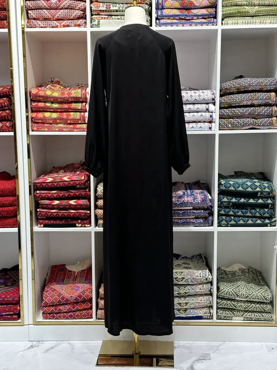 Black umra abaya for women displayed on mannequin with colorful folded fabrics in background