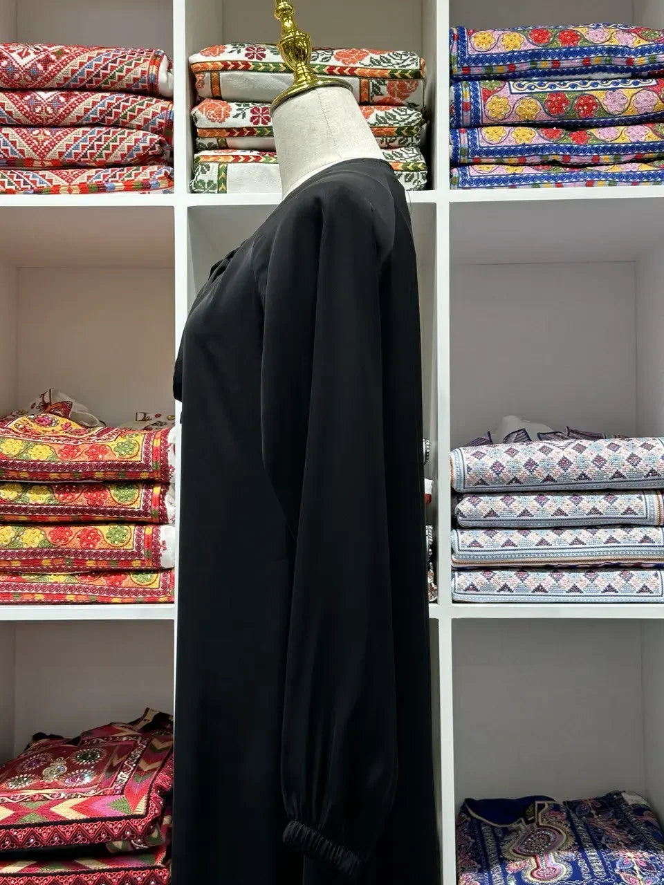 Black umra abaya for women displayed on a mannequin with colorful fabric shelves in the background