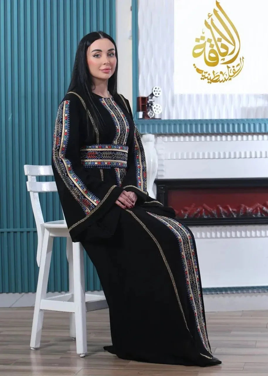 Woman wearing a black embroidered Palestinian kaftan with multicolor traditional designs sitting on a white chair indoors