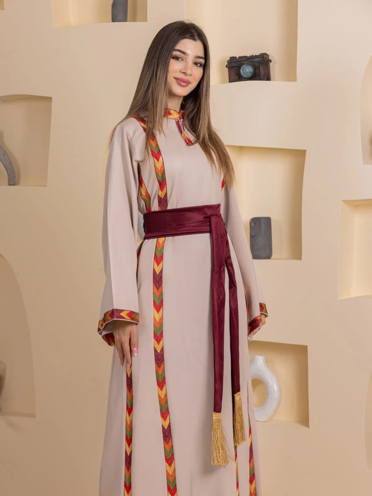 Palestinian Elegance embroidered beige thobe with maroon sash and colorful chevron trim on model Embroiderd Thoub Al-Manajel