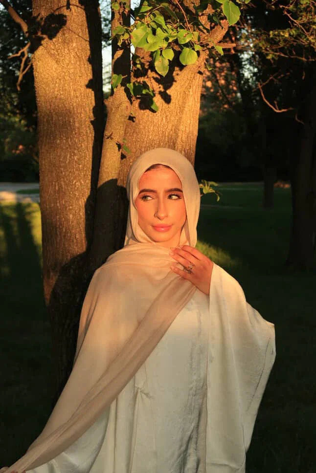 Model wearing New Modal Hijab by Palestinian Elegance in beige drape and white outfit standing by a tree in golden hour light