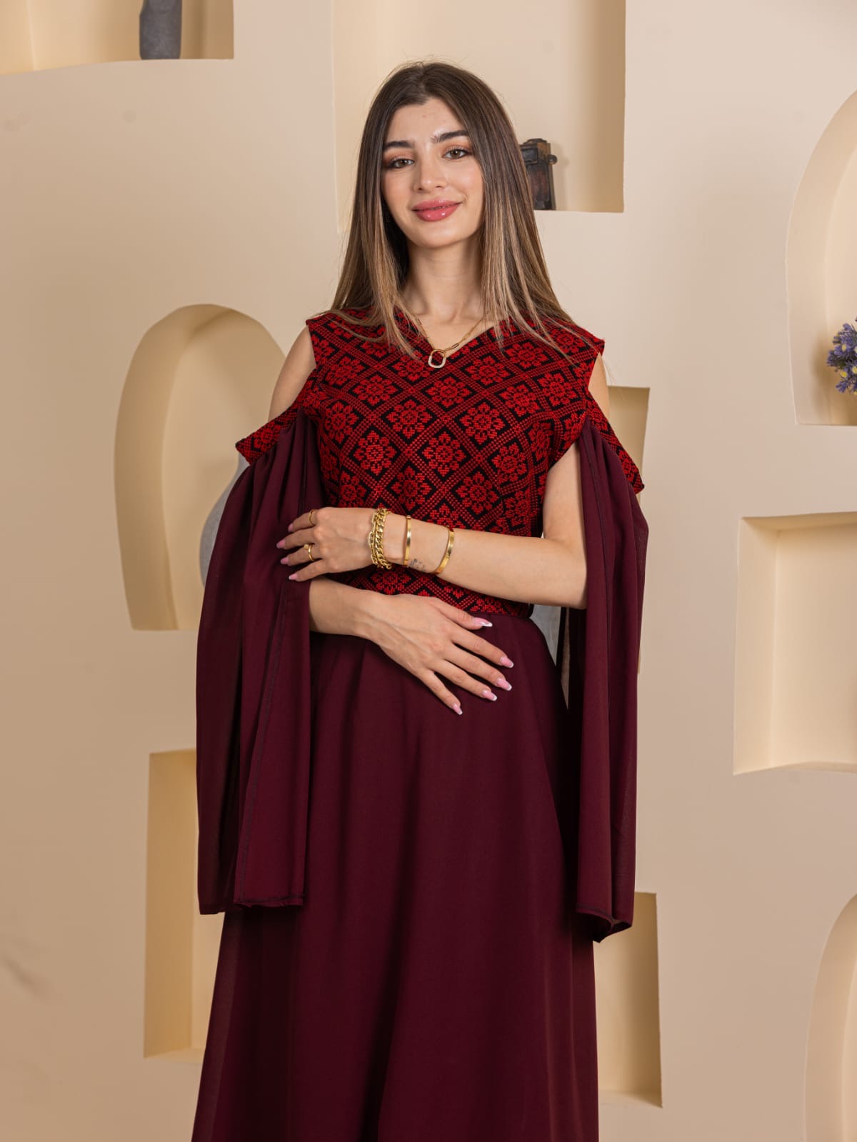 Model wearing Palestinian Essence Dress burgundy gown with traditional red embroidered bodice and draped cold shoulder sleeves