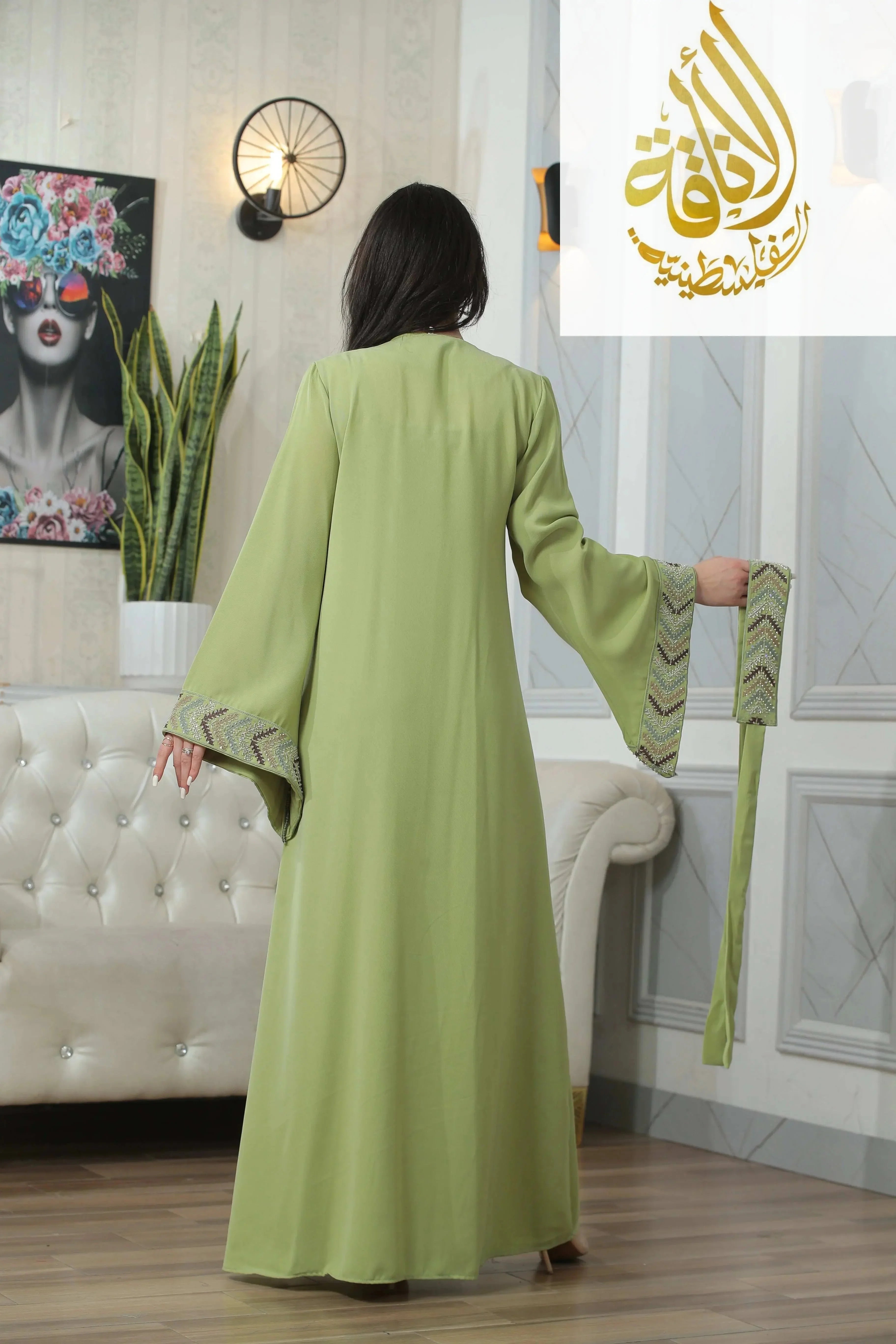 Back view of a woman wearing a green embroidered beaded thobe dress for women holding a matching belt indoors
