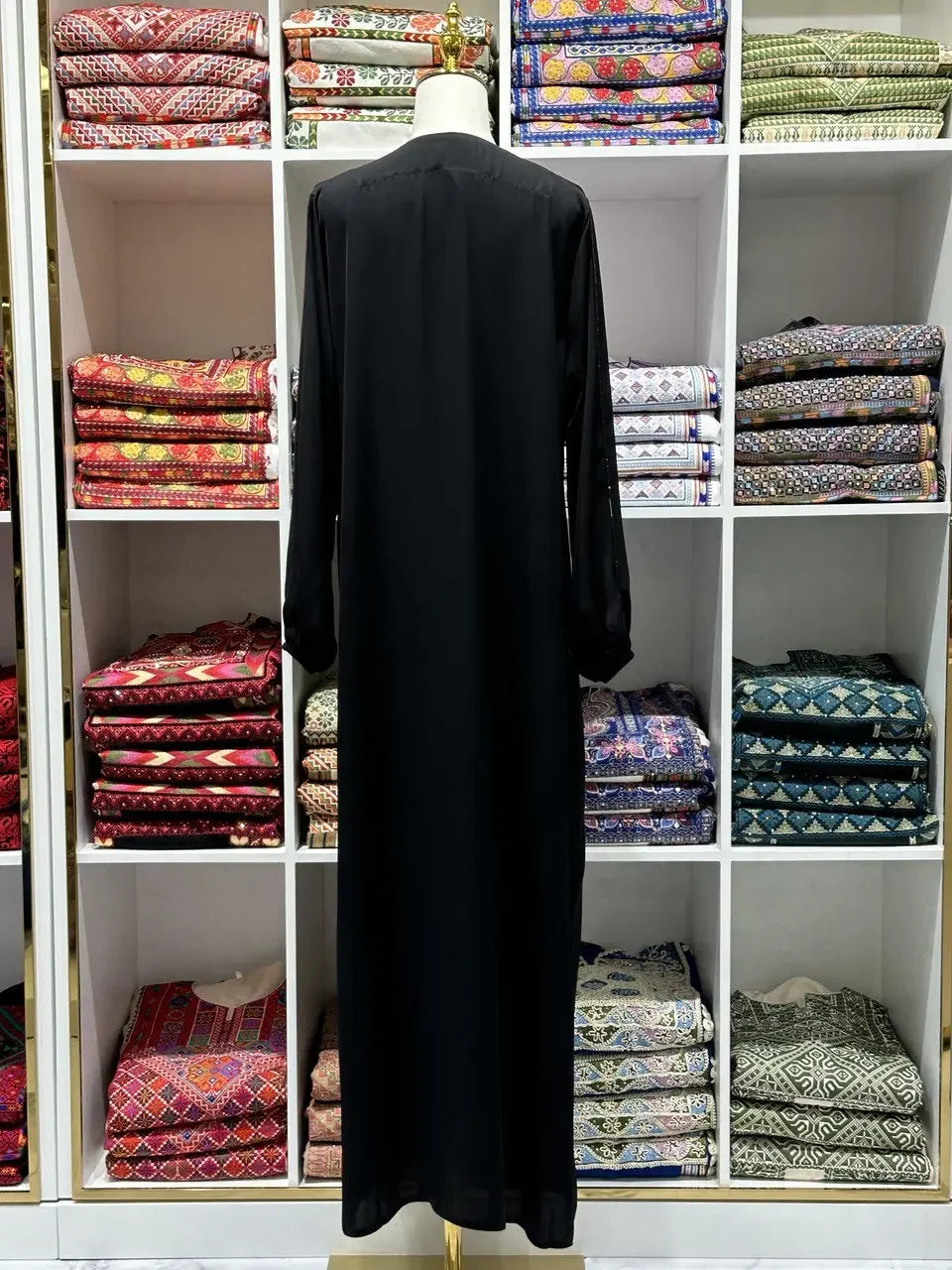 Black abaya simple design displayed on a mannequin in front of colorful folded fabrics by Palestinian Elegance