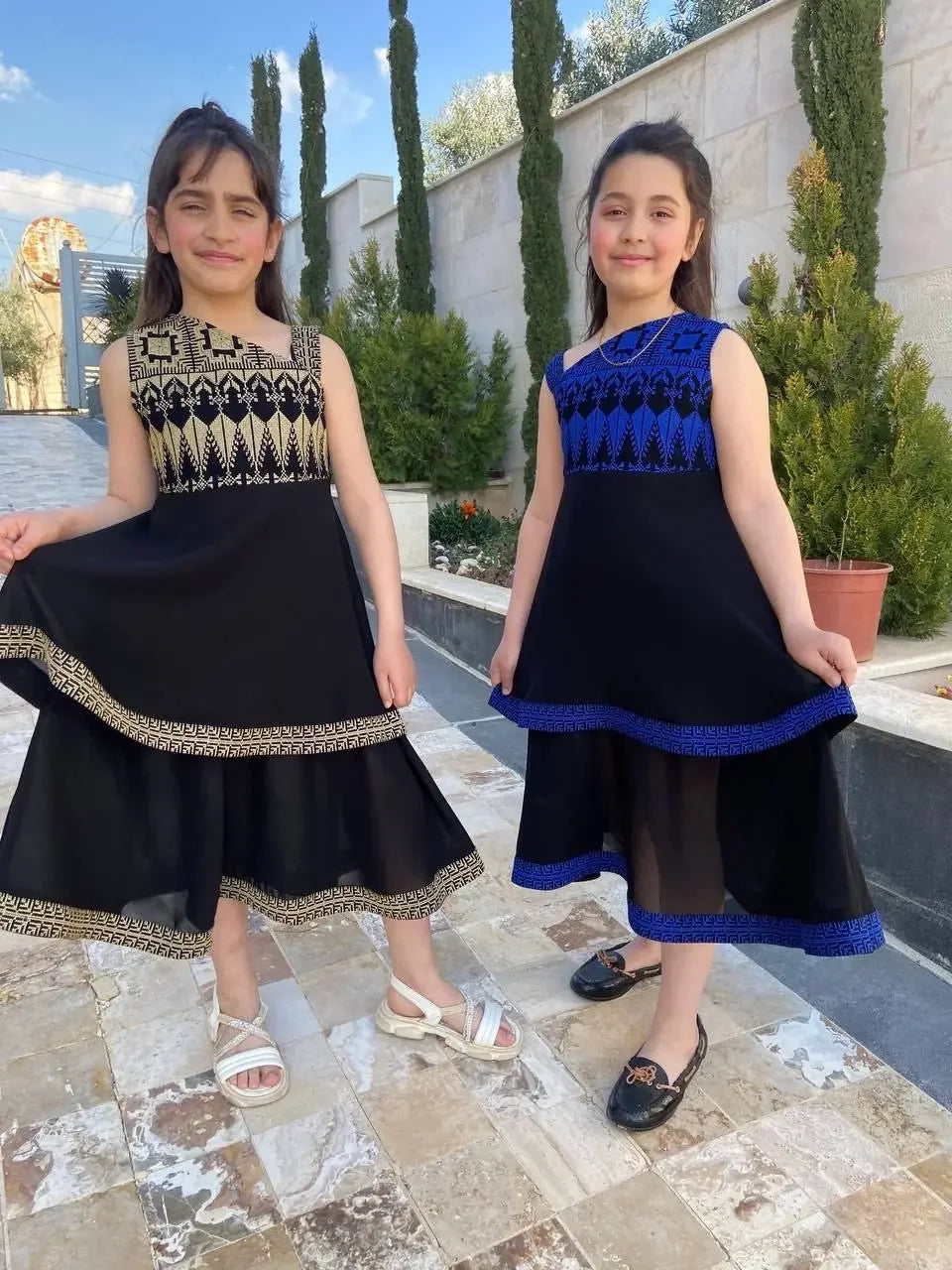 Two girls wearing embroidered dress for kids by Palestinian Elegance in black with gold and blue details outdoors