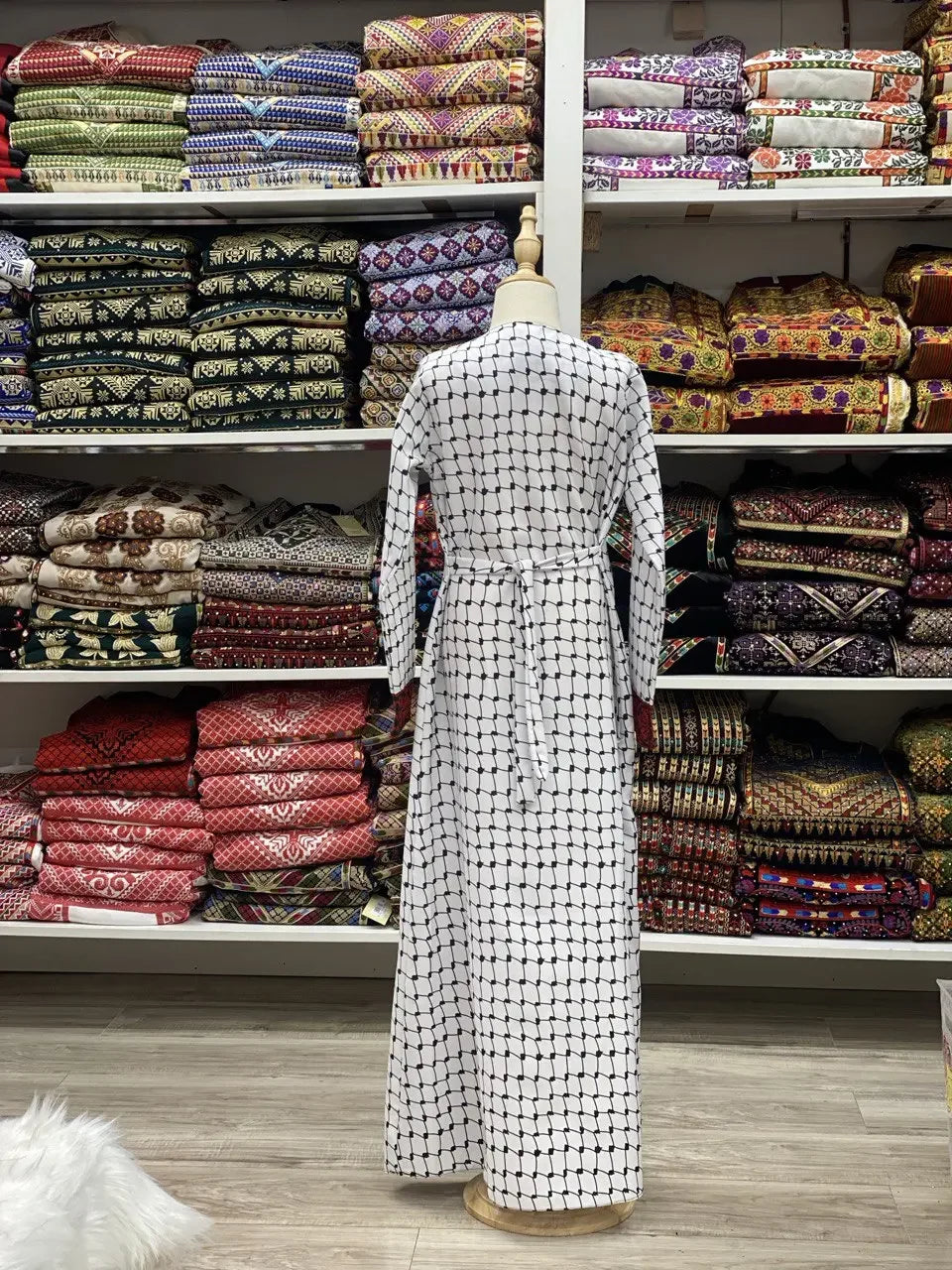 Kids embroidered Palestinian dress displayed on a mannequin in a store with folded traditional textiles in the background