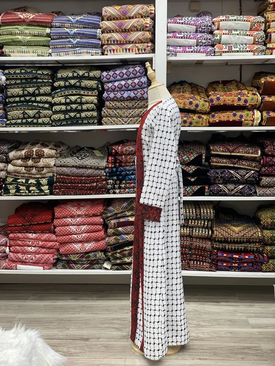 Kids embroidered Palestinian dress in white with traditional red and black stitching displayed on a mannequin in front of colorful folded fabrics