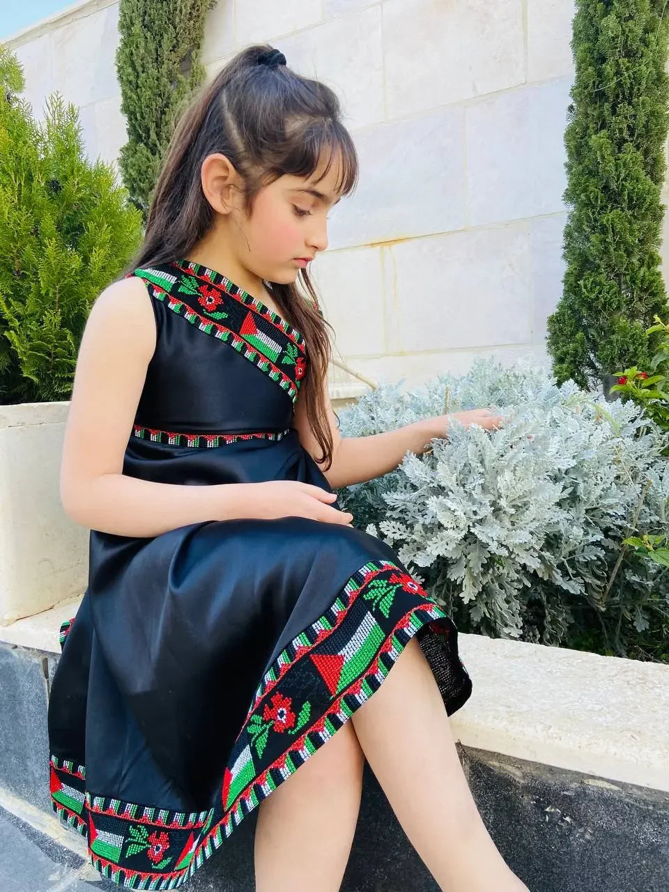 Girl wearing black kids Palestine dress with traditional red and green embroidery sitting by garden plants