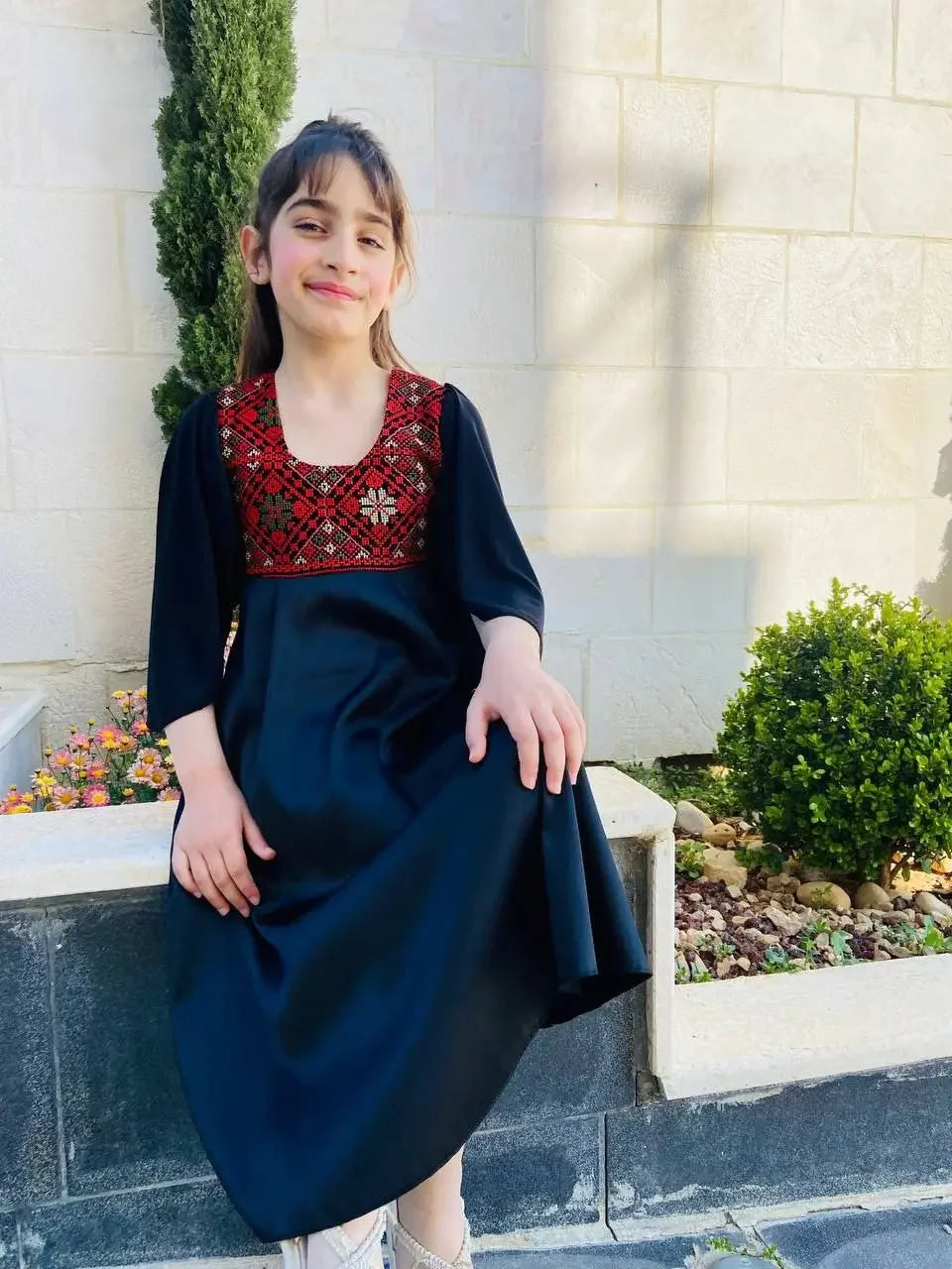 Girl wearing black kids tatreez dress with traditional red embroidery sitting outdoors by garden
