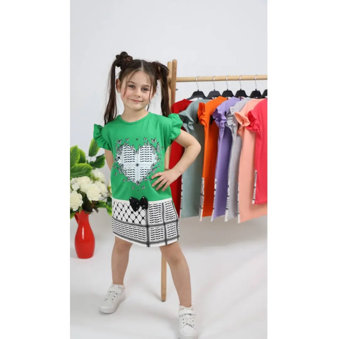 Girl wearing green kuffiyeh print dress with heart design standing in front of colorful dresses on rack
