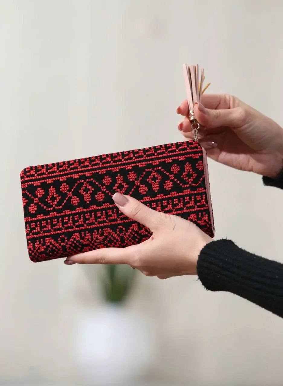 Hand holding a Palestinian embroidered wallet with red and black traditional patterns and tassel accessory