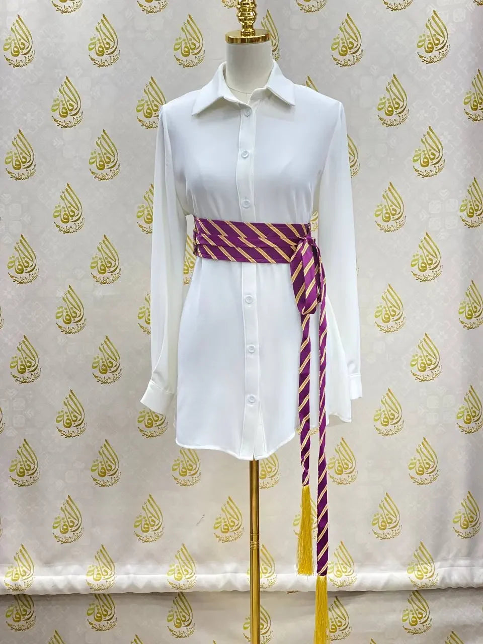 Embroidered kashmir belt in purple and gold stripes tied around a white shirt dress by Palestinian Elegance