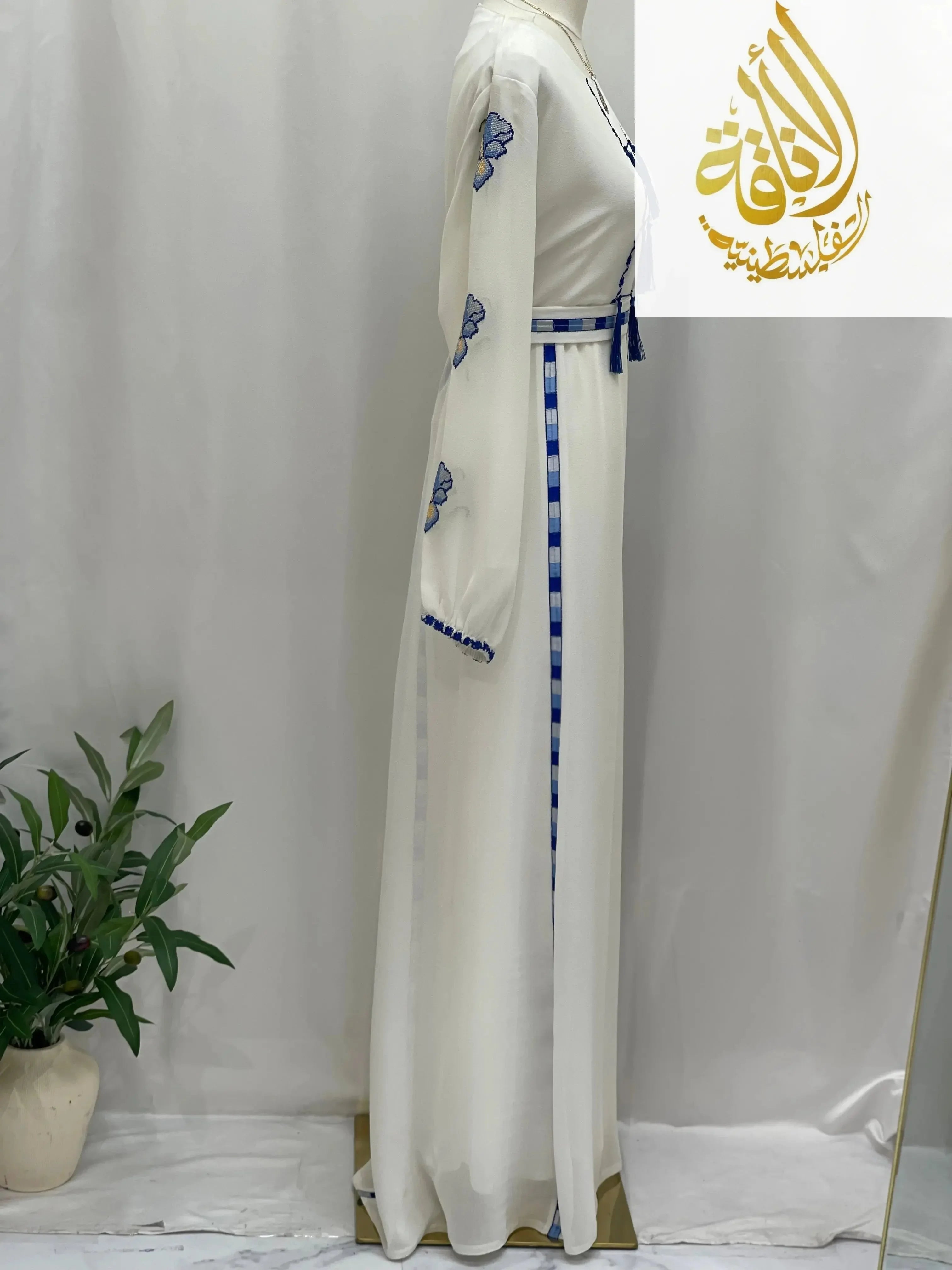 White Palestinian embroidered dress with blue floral designs and tassels by Palestinian Elegance