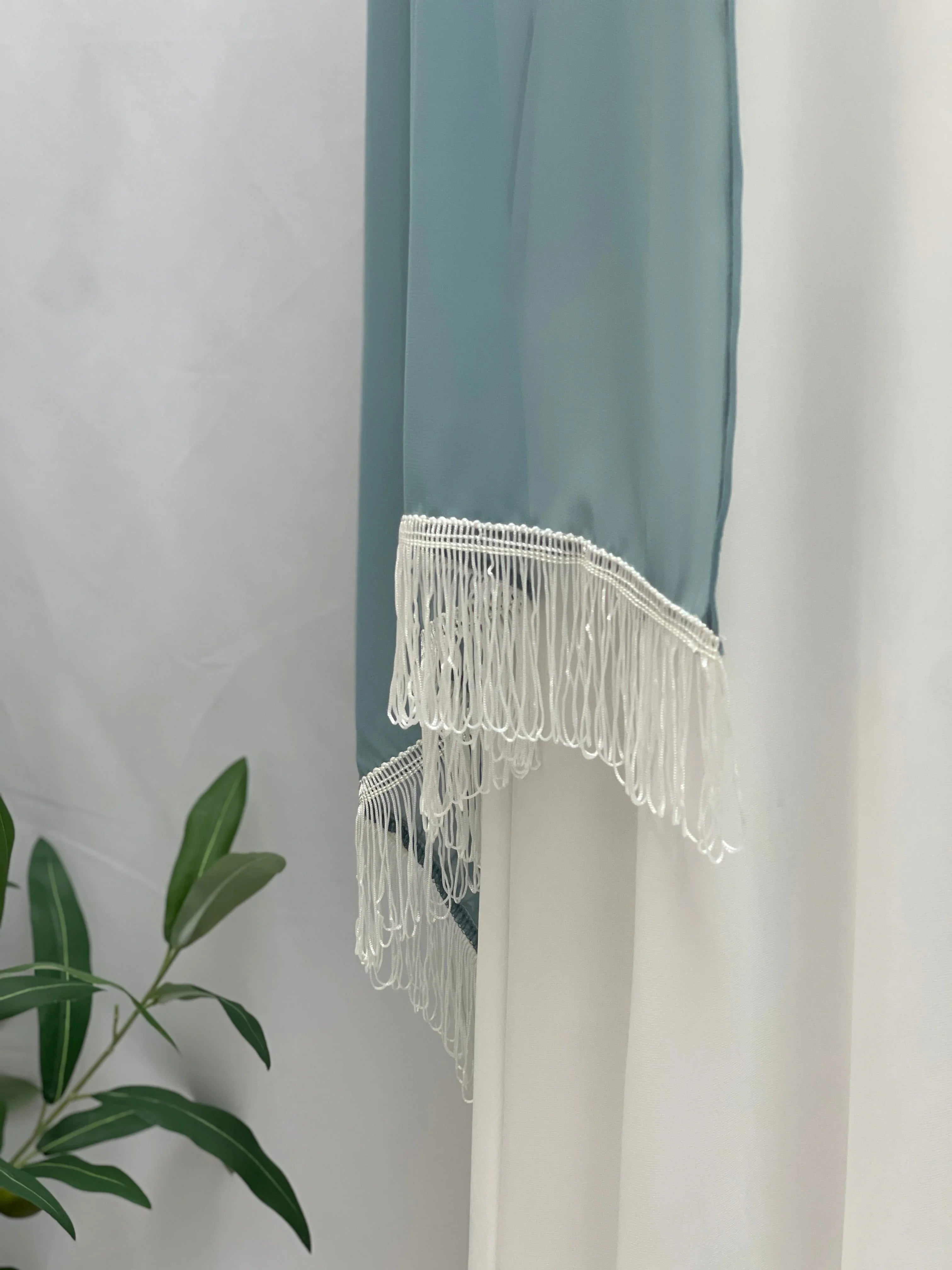New Khirka With White tassel teal Palestinian Elegance fringe hem draped against white curtain and green plant