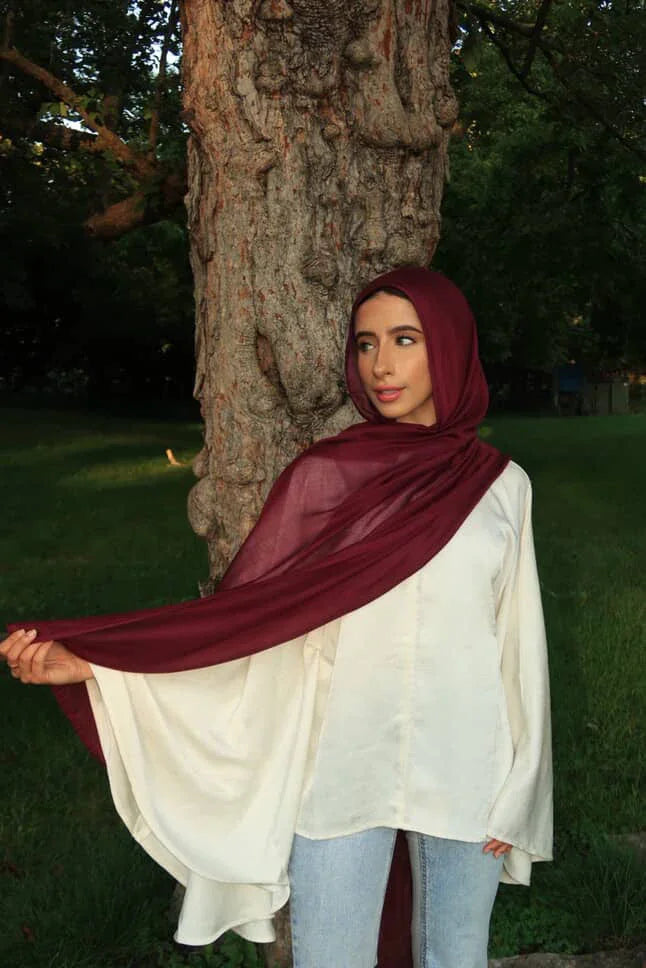 Palestinian Elegance New Modal Hijab in burgundy on model with white flowy blouse and jeans by a tree