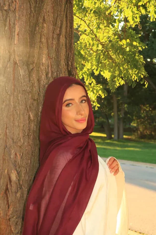 Palestinian Elegance model wearing New Modal Hijab in deep burgundy leaning against tree in sunlit park