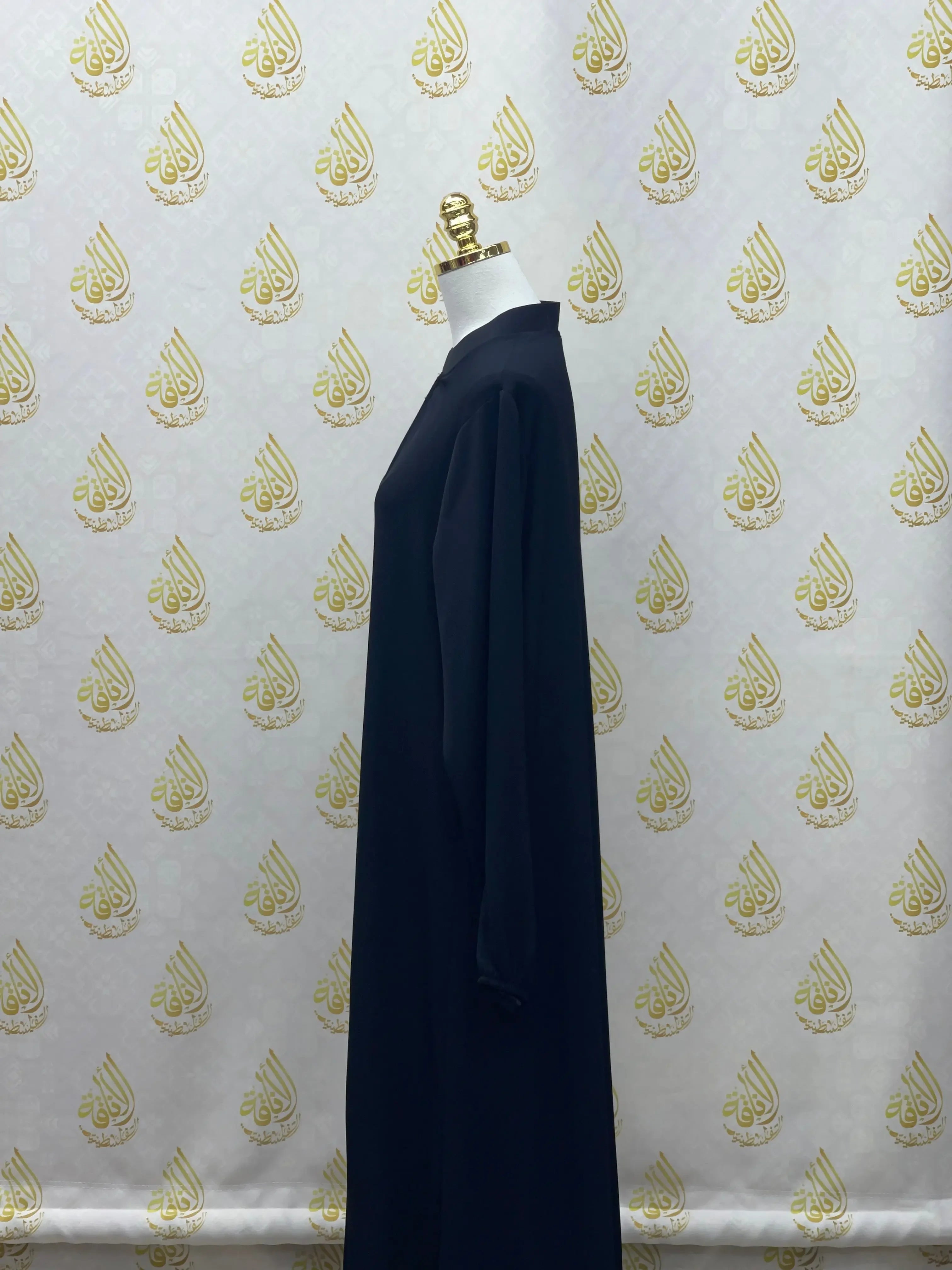 Plain black abaya displayed on mannequin by Palestinian Elegance in side view against patterned backdrop
